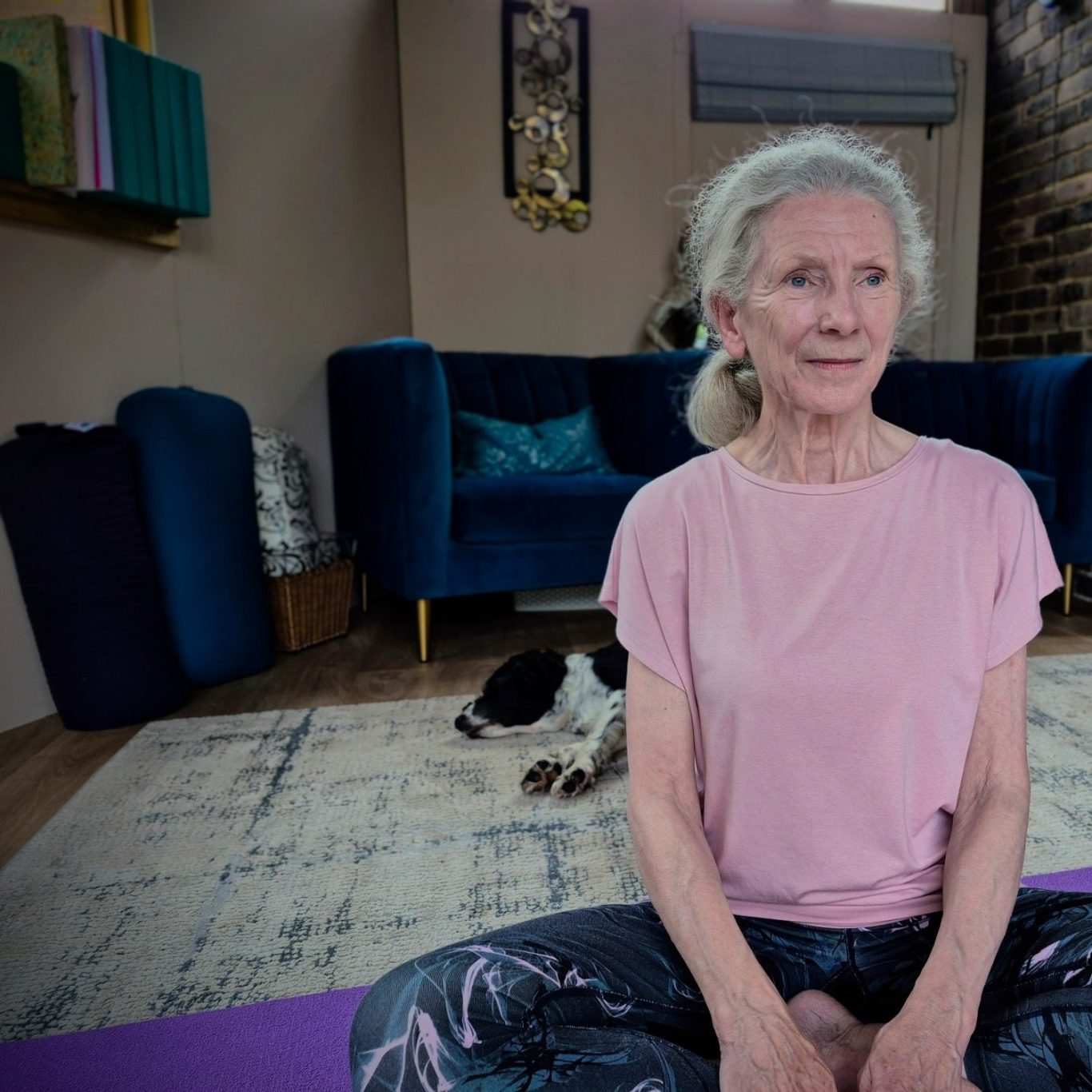 Elderly woman seated in a yoga pose, smiling, with a dog resting nearby.